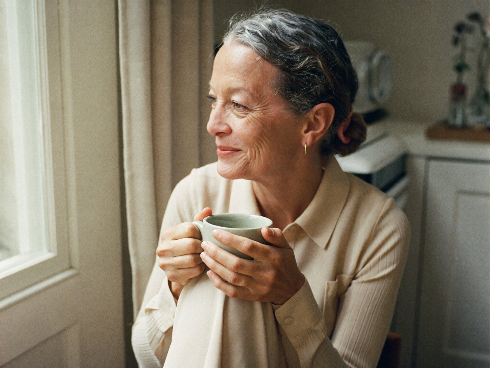 A woman in soft morning light by a window, calm and reflective, a mug in hand.
