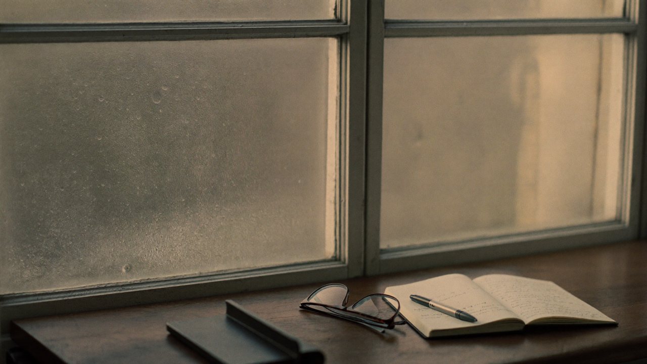 A window beaded with morning condensation beside an open notebook and reading glasses.